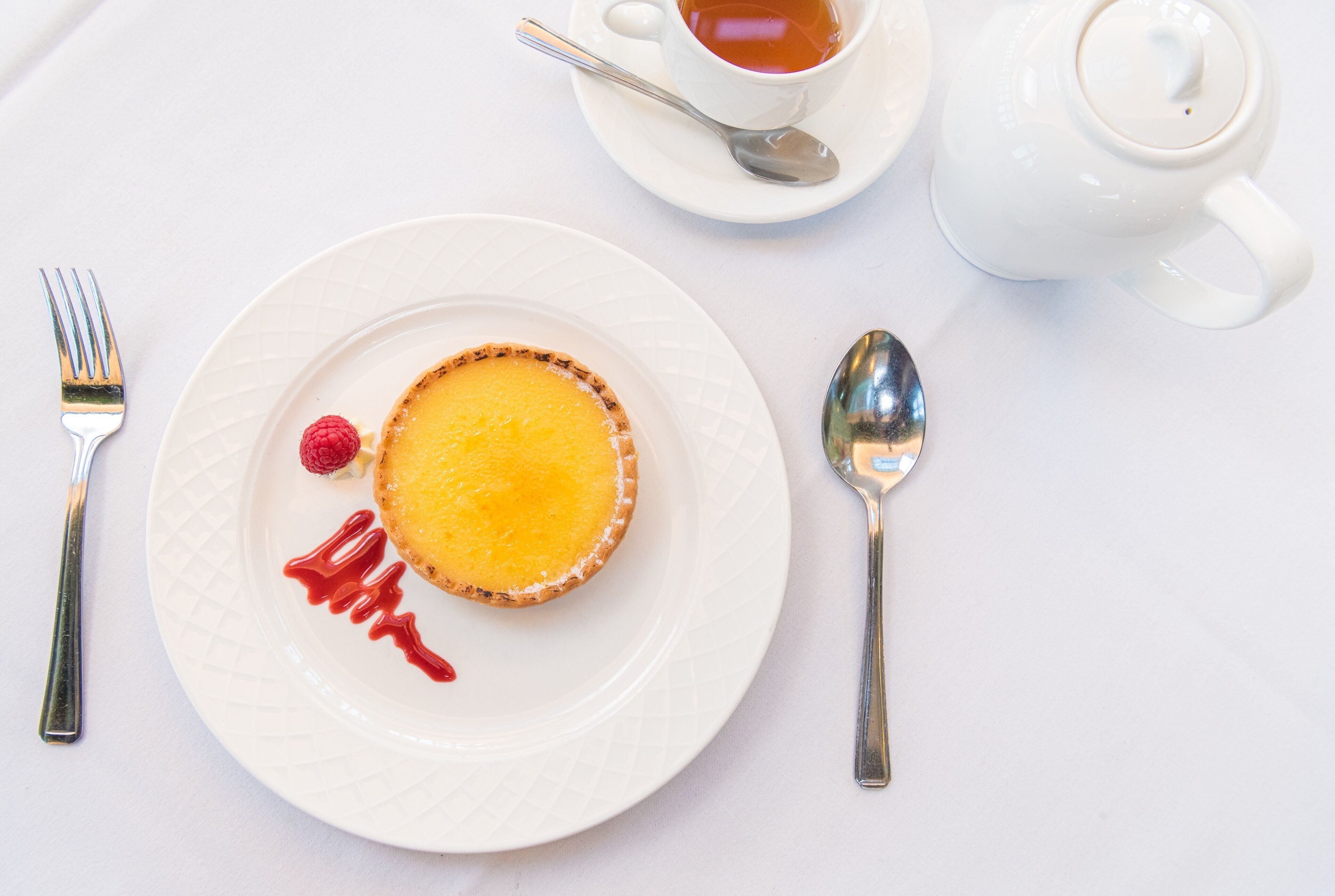 A dessert plate with a tart, a cup of tea, and a teapot on a white table.