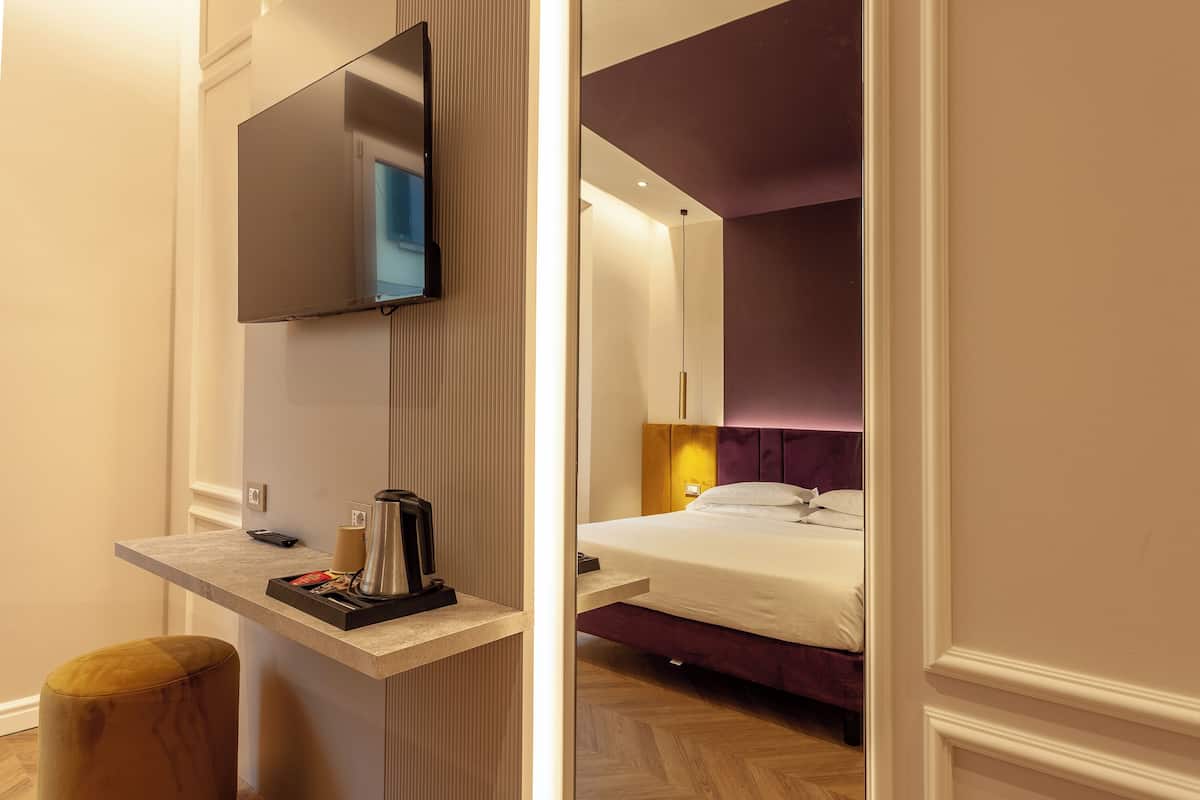 Camera d'albergo moderna con TV a schermo piatto fissata al muro, un piccolo comodino con una macchina per il caffè e un letto con testiera viola.