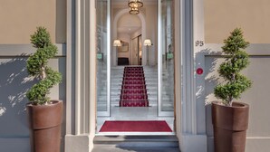 Entrada de hotel con alfombra roja, escalones de mármol y dos plantas en macetas.