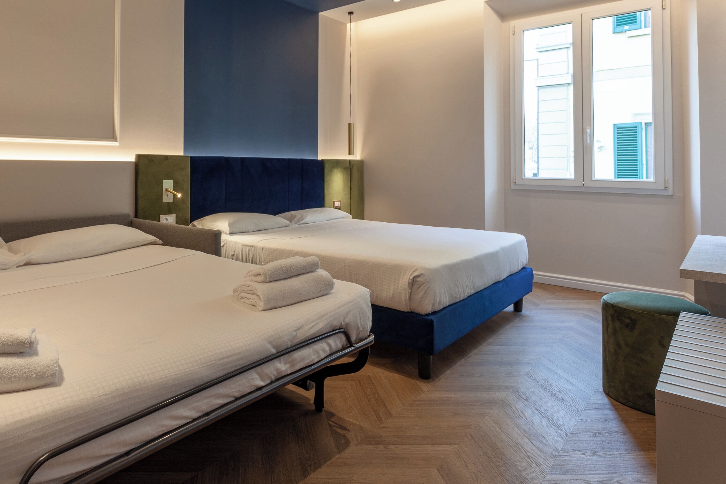 A hotel room with two beds, a green ottoman, and a desk.