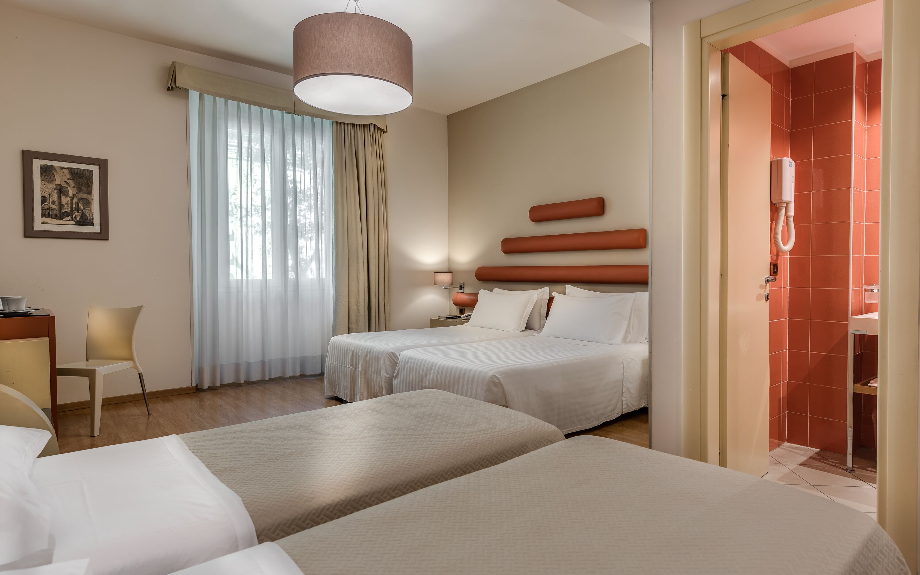 A hotel room with two beds, a desk, a chair, a window with curtains, and a bathroom visible through an open door.