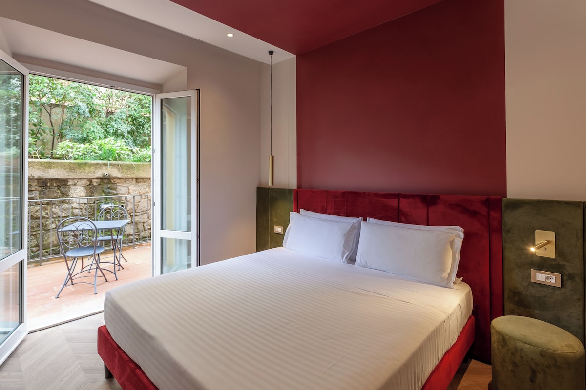 A bedroom with a large bed, a red headboard, a small green ottoman, and a balcony with a table and chairs.