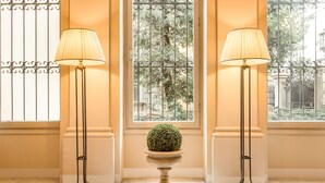Dos lámparas de pie con pantallas, una planta pequeña en un pedestal y una ventana que ofrece vistas a árboles y edificios.