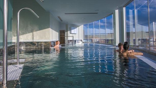 Una piscina interior con paredes de vidrio transparente, techo curvo y vista al cielo.
