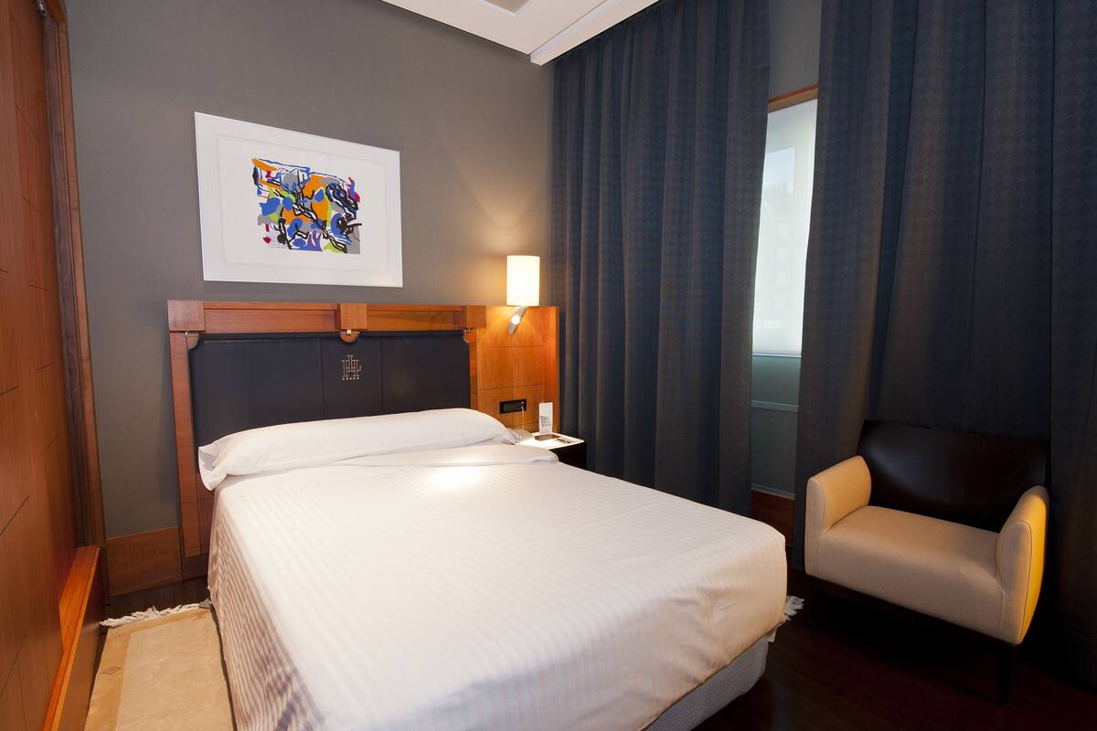 A hotel room with a bed, a chair, a nightstand, a lamp, and a framed artwork on the wall.