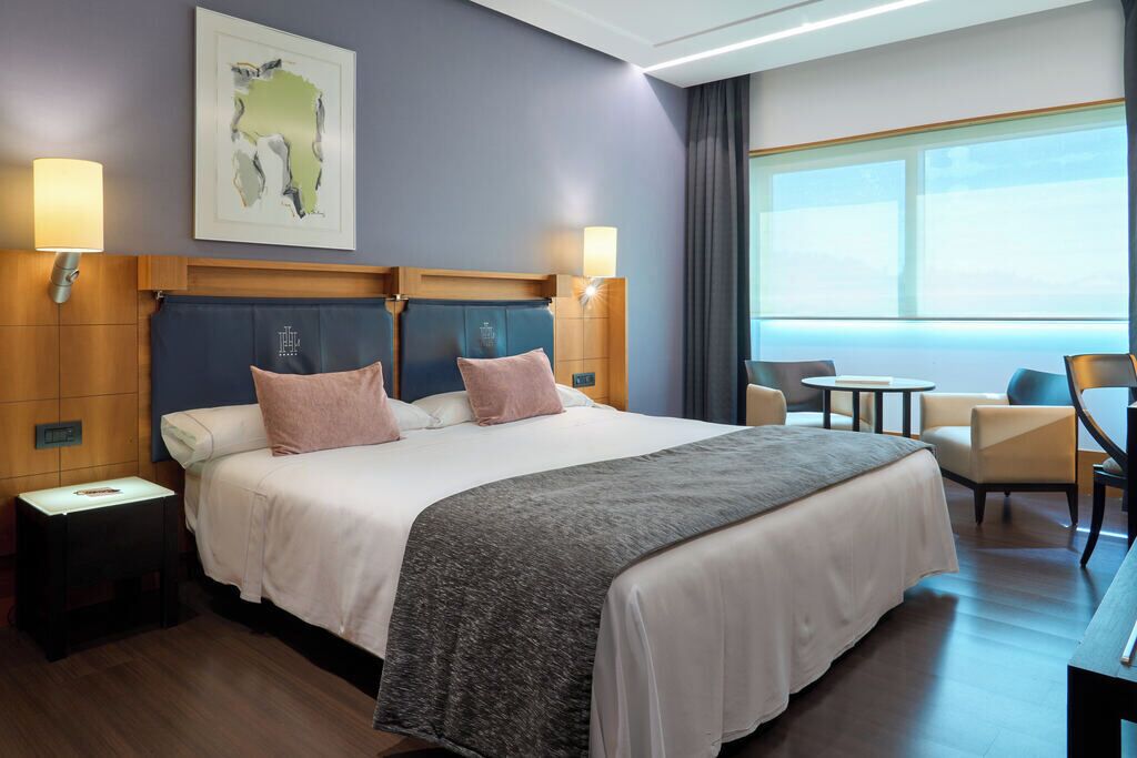 A hotel room with a large bed, two bedside tables, a desk with a chair, and a view of the outdoors.