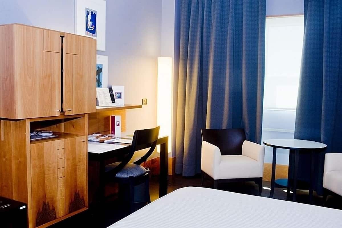 A hotel room with a bed, a desk with a chair, a small table, and a wardrobe.