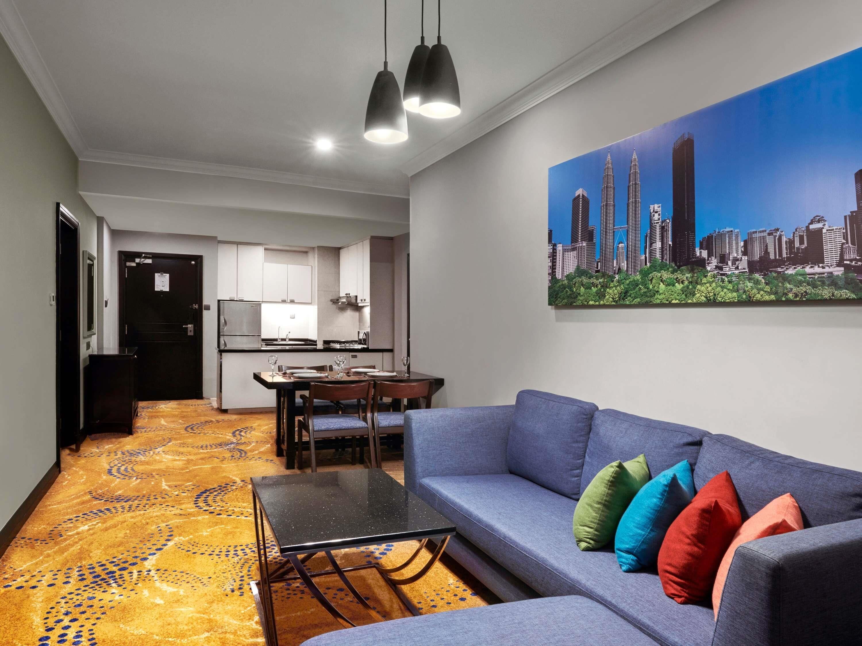A modern hotel room with a dining area, a sofa with colorful cushions, and a cityscape painting on the wall.