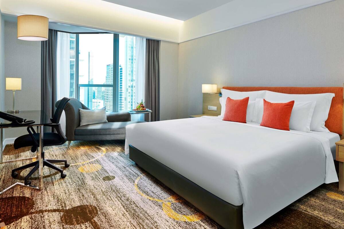 A hotel room with a large bed, a desk, a chair, and a view of the city through large windows.