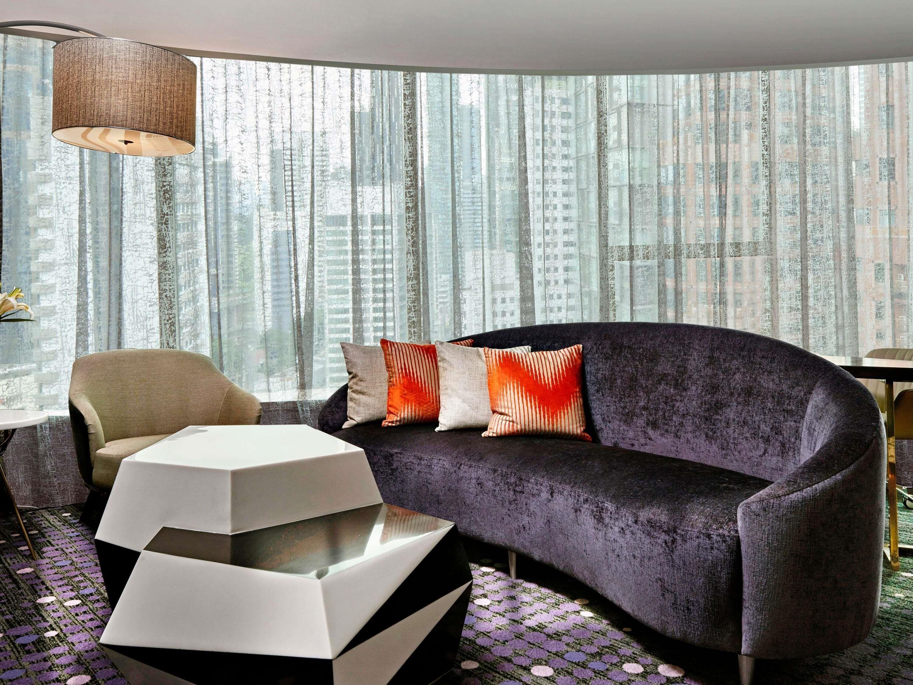 A modern living room with a sofa, a chair, a coffee table, and a view of tall buildings through sheer curtains.
