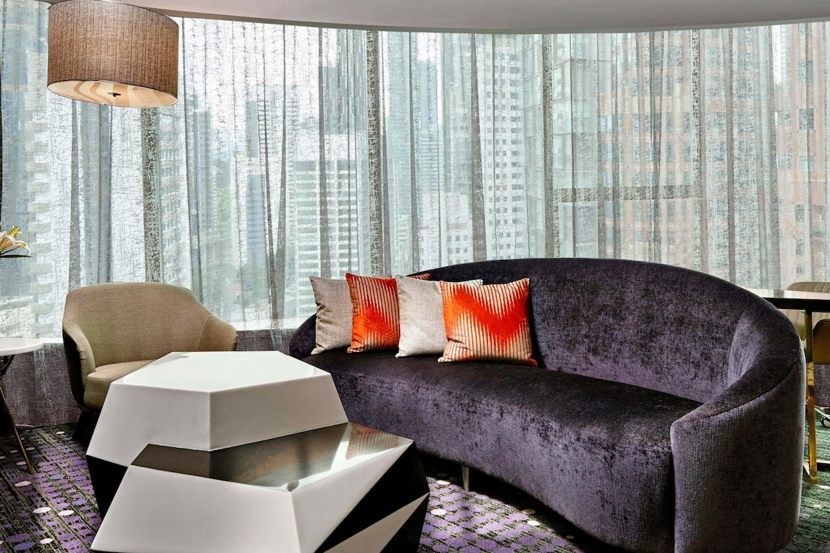 A modern living room with a sofa, a chair, a coffee table, and a view of tall buildings through sheer curtains.