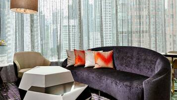 A modern living room with a sofa, a chair, a coffee table, and a view of tall buildings through sheer curtains.