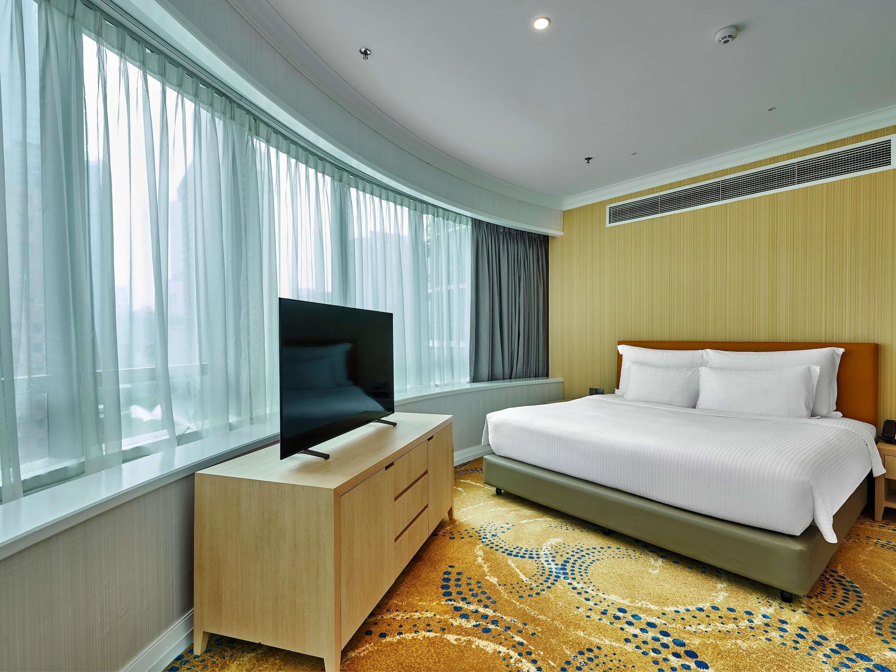 A hotel room with a bed, a TV on a wooden dresser, a window with sheer curtains, and a patterned carpet.