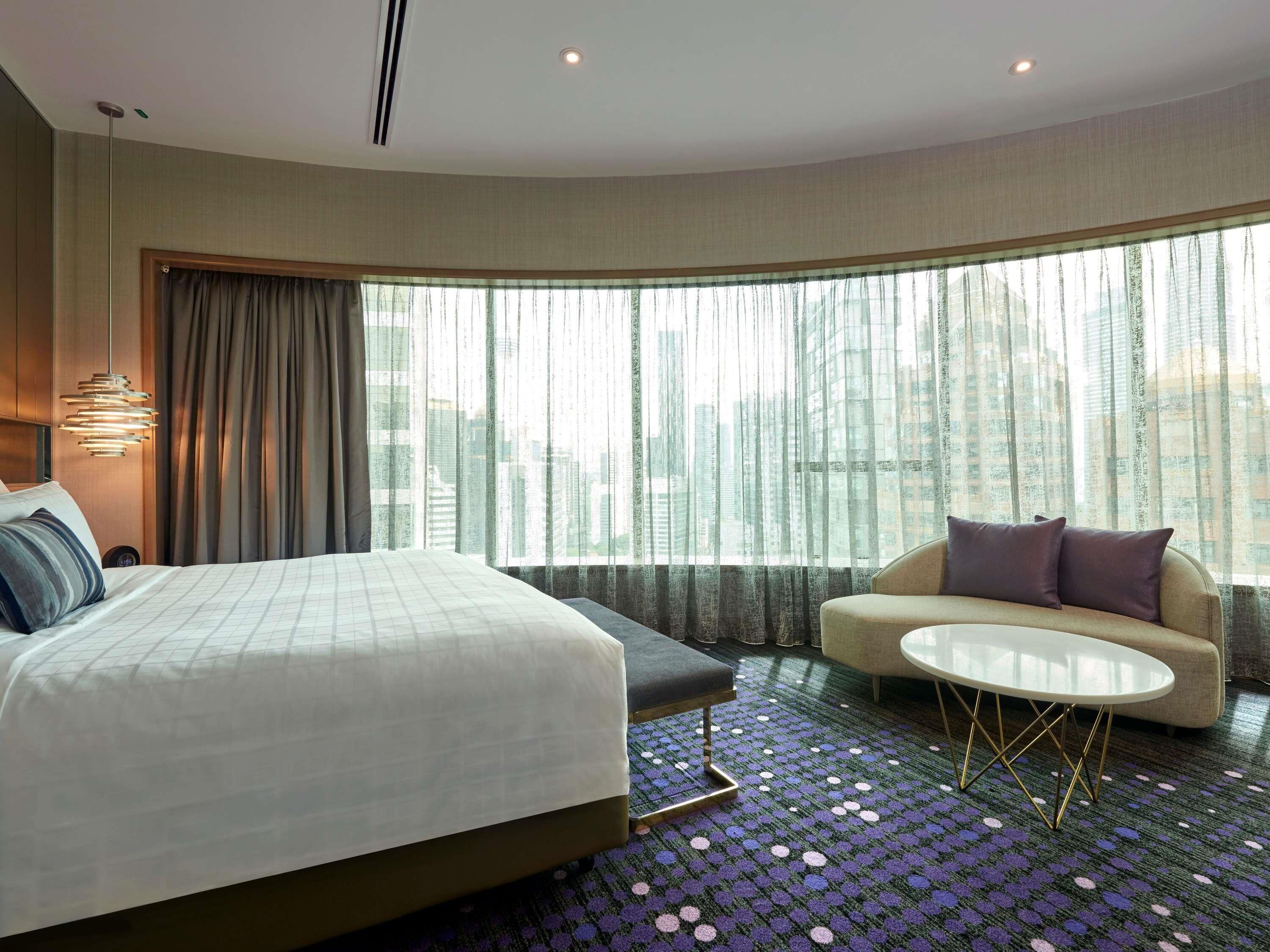 A hotel room with a bed, a sofa, a small table, and a view of the city through large windows.