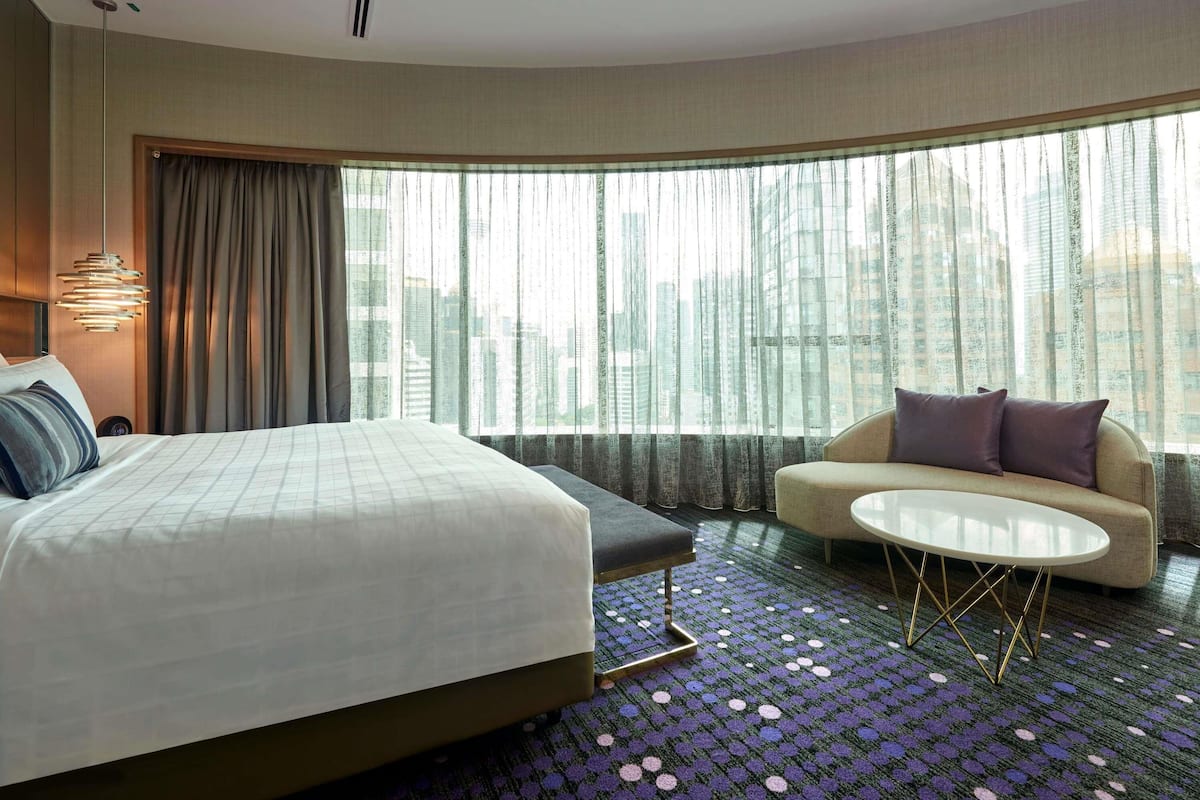 A hotel room with a bed, a sofa, a small table, and a view of the city through large windows.