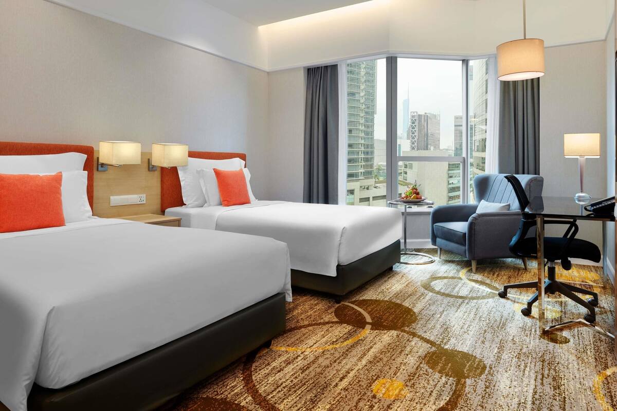 A hotel room with two beds, a desk, a chair, a large window with a city view, and a patterned carpet.