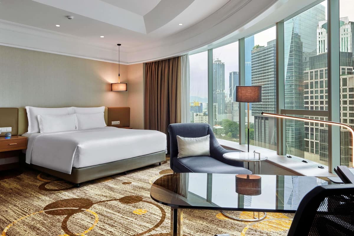 A hotel room with a bed, a chair, a desk, and a large window offering a city view.