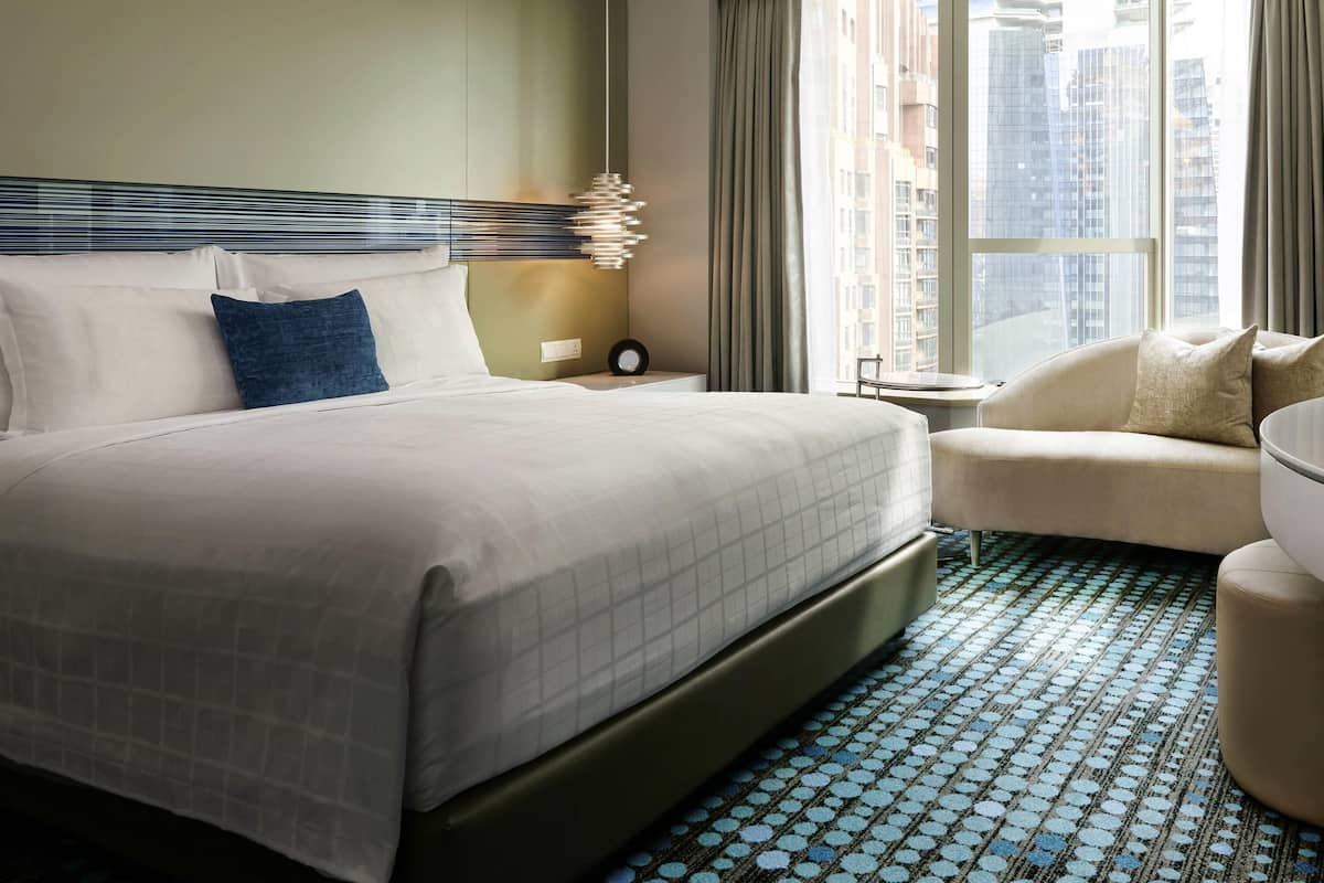 A hotel room with a large bed, a seating area, a window with a city view, and a patterned floor.