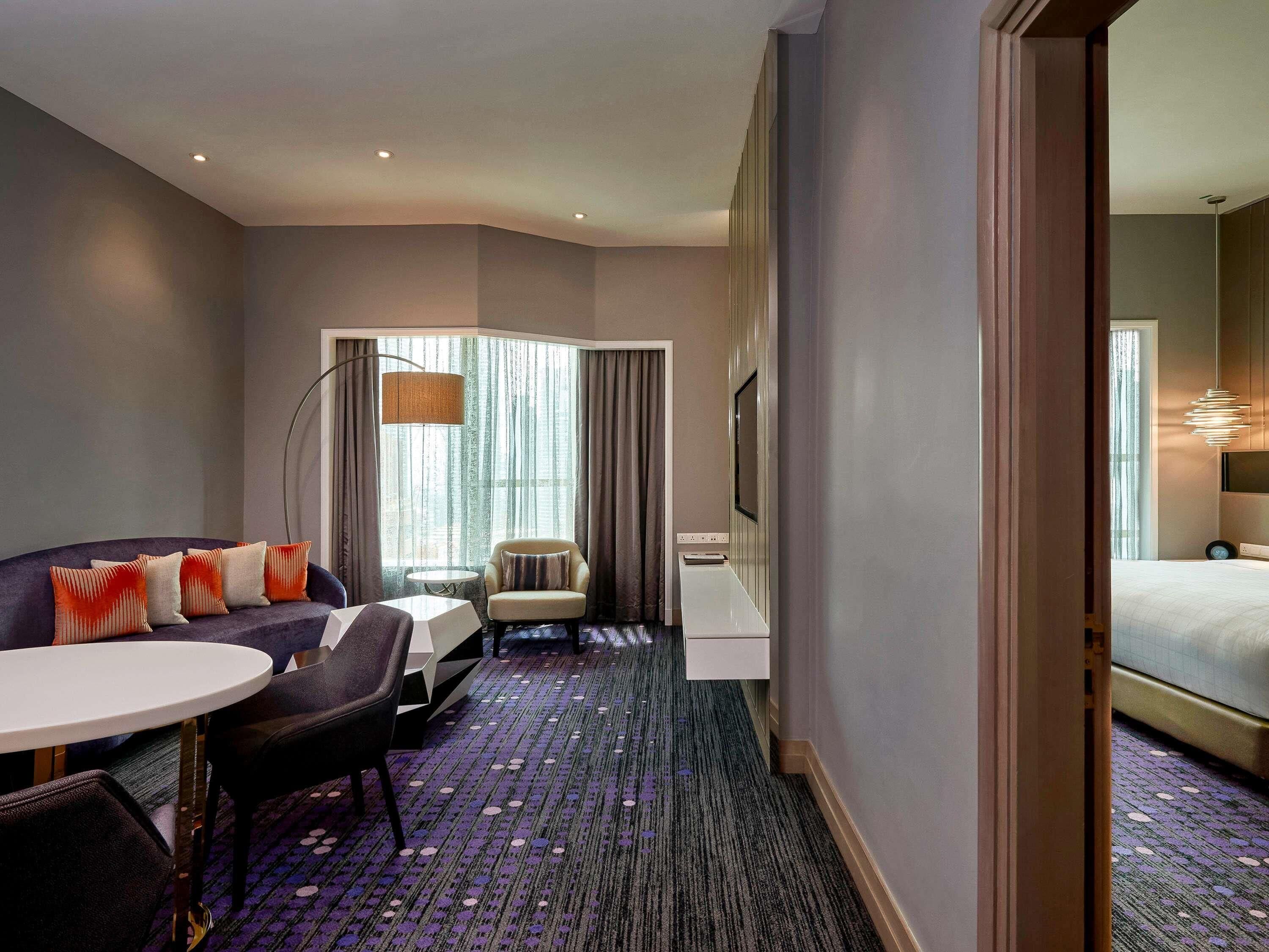 Quarto de hotel com cama, sofá, escrivaninha, cadeira, mesinha e uma grande janela com cortinas.