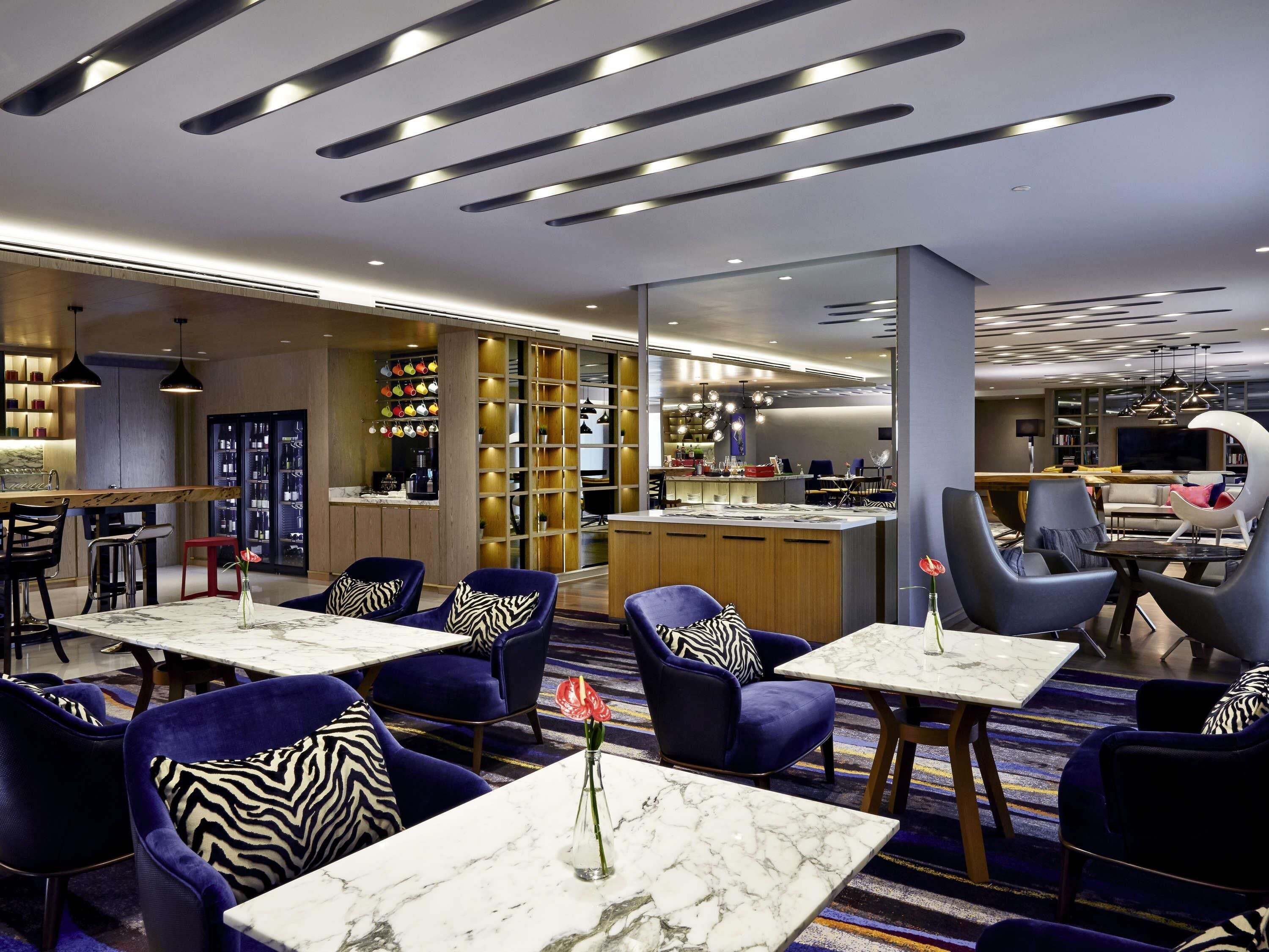 Un hall d’hôtel moderne avec un bar, des sièges dotés de chaises bleues et de coussins à motifs zébrés, et une cuisine visible au fond.