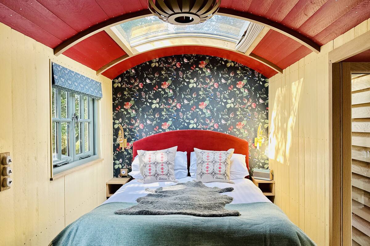 Un dormitorio con cabecera roja, papel tapiz floral y tragaluz sobre la cama.