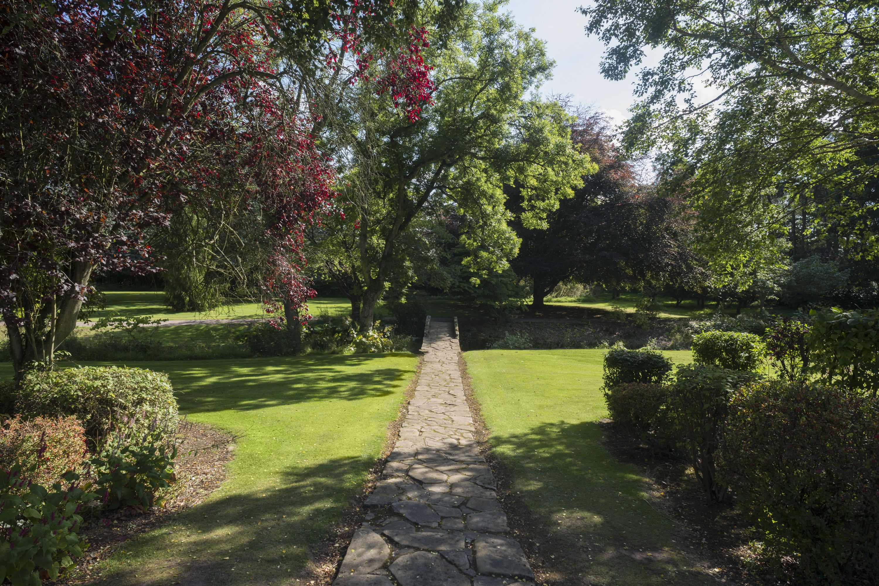 A stone pathway leading through a well-maintained garden with various trees and shrubs.