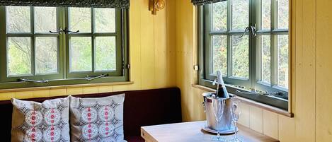 A cozy room with a wooden bench, a small table with a wine glass, and two windows with curtains.