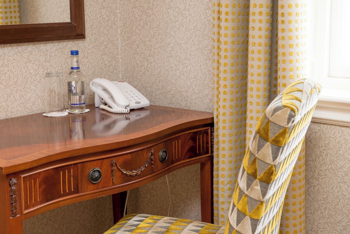 Een houten bureau met een telefoon, een fles water en een stoel met een gestreept kussen.