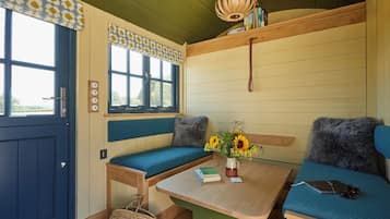 A cozy room with a blue door, a bench with a blue cushion, a table with a book and a mug, and a shelf with books and a lamp.