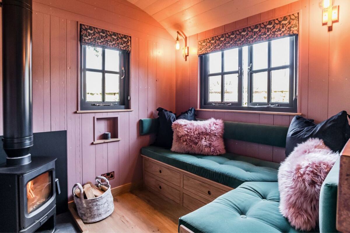 Interior acogedor de una cabaña con estufa de leña, un banco verde con cojines y dos ventanas con marcos decorativos.