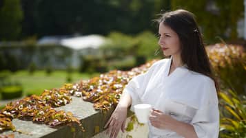 A person in a white robe holding a cup, standing by a wall with vines.