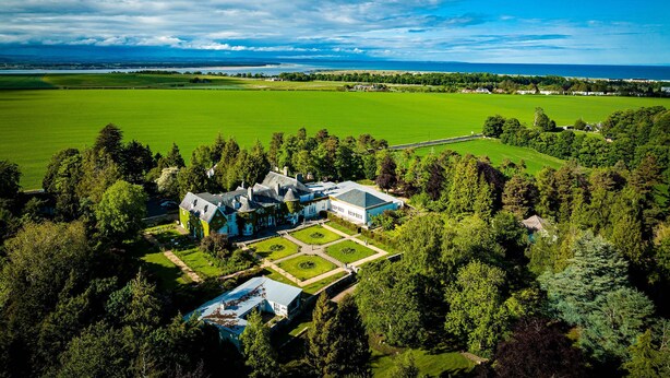 An aerial view of a large estate with expansive green fields, a formal garden, and a main building with multiple sections.
