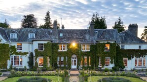 A large, historic building with a symmetrical facade, a central entrance, and a well-maintained garden with a stone pathway leading to the entrance.