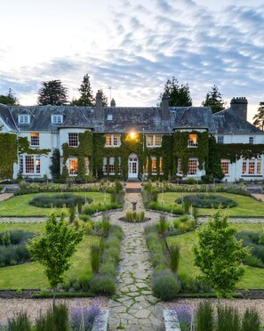 A large, historic building with a symmetrical facade, a central entrance, and a well-maintained garden with a stone pathway leading to the entrance.