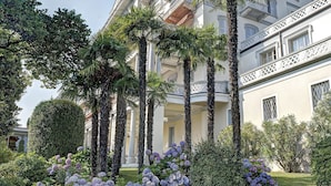 Un hôtel à plusieurs étages, doté d’une façade symétrique, de balcons et d’un jardin bien entretenu, agrémenté de haies et de plantes à fleurs.