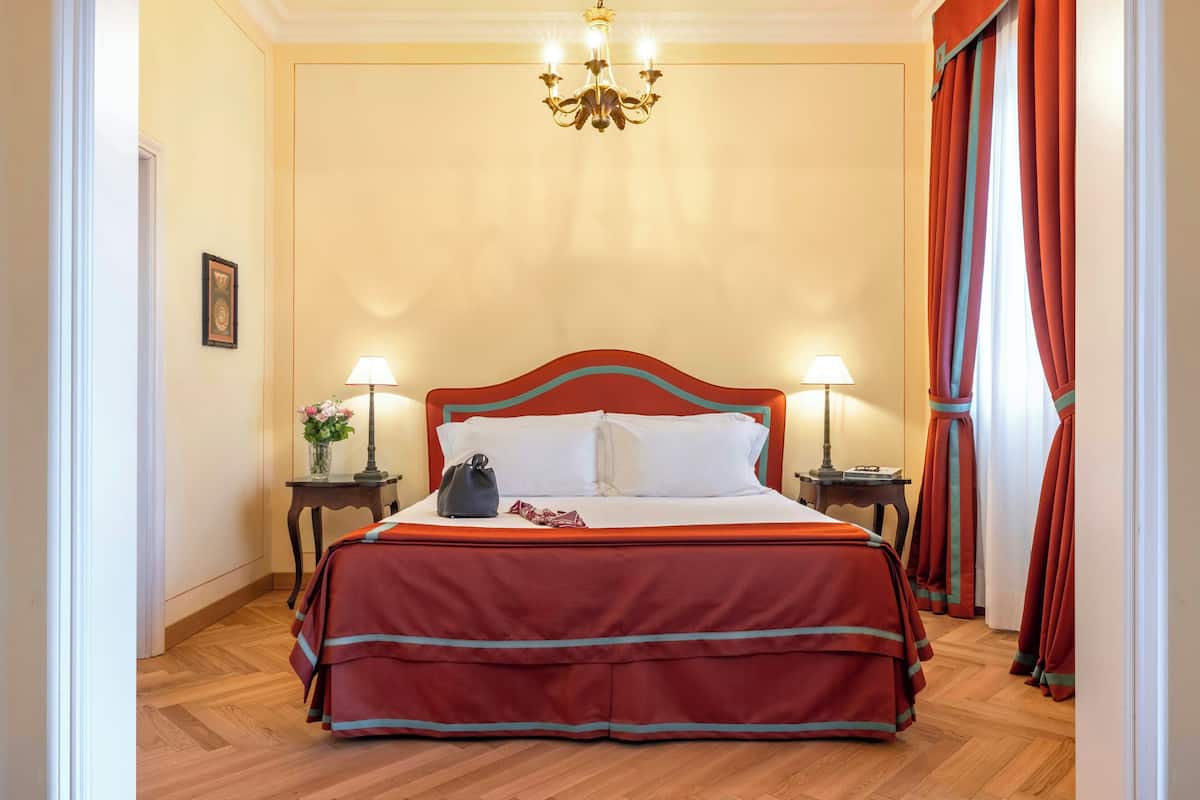 Hotelkamer met een groot bed, twee nachtkastjes, een kroonluchter en rode gordijnen.