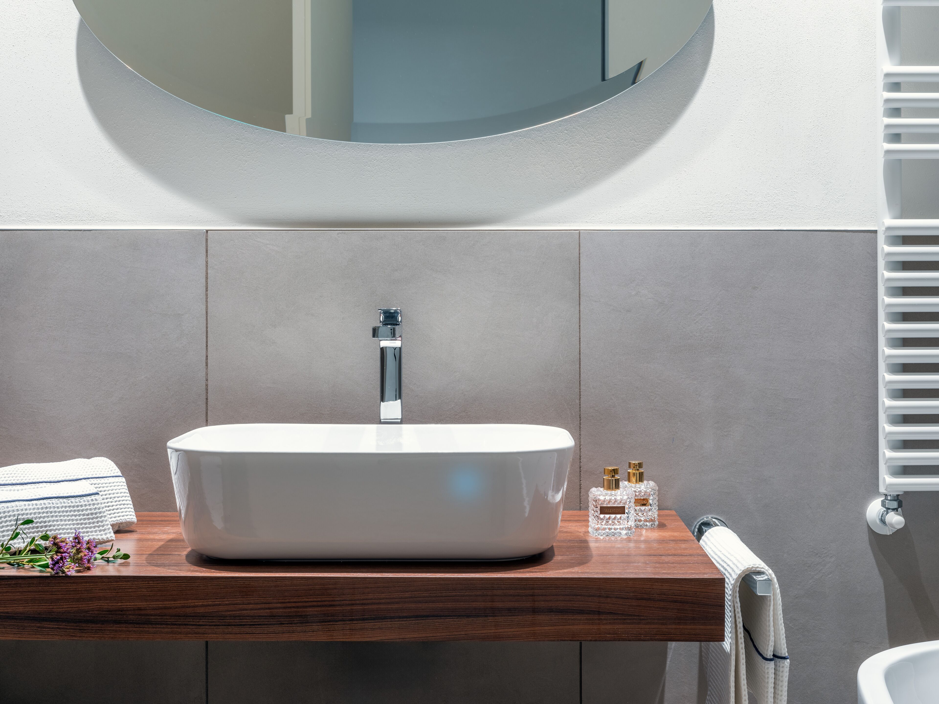 Un bagno moderno con un lavandino ovale bianco, uno specchio rotondo e un piano d'appoggio in legno.