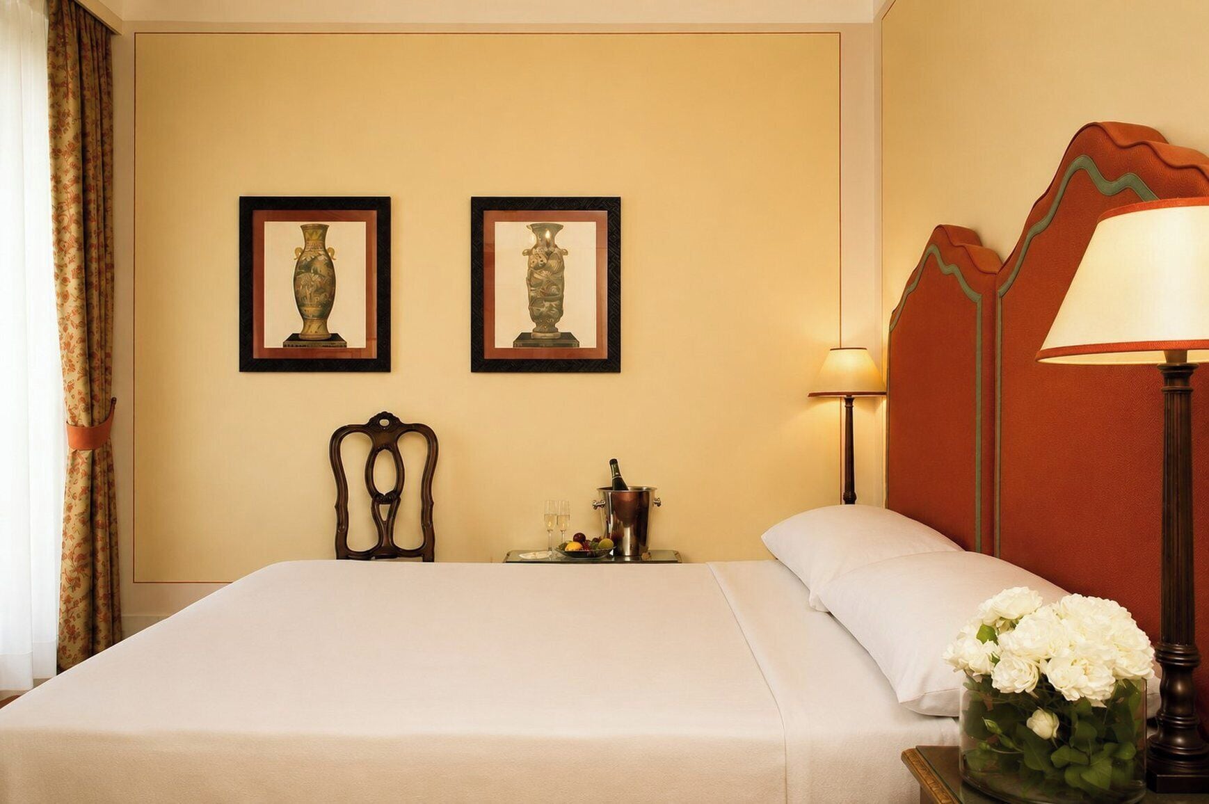 A hotel room with a bed, two framed vases, a nightstand with a bottle and glasses, a lamp, and a vase of flowers.