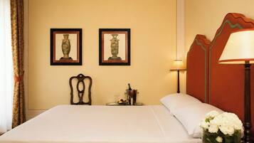 A hotel room with a bed, two framed vases, a nightstand with a bottle and glasses, a lamp, and a vase of flowers.