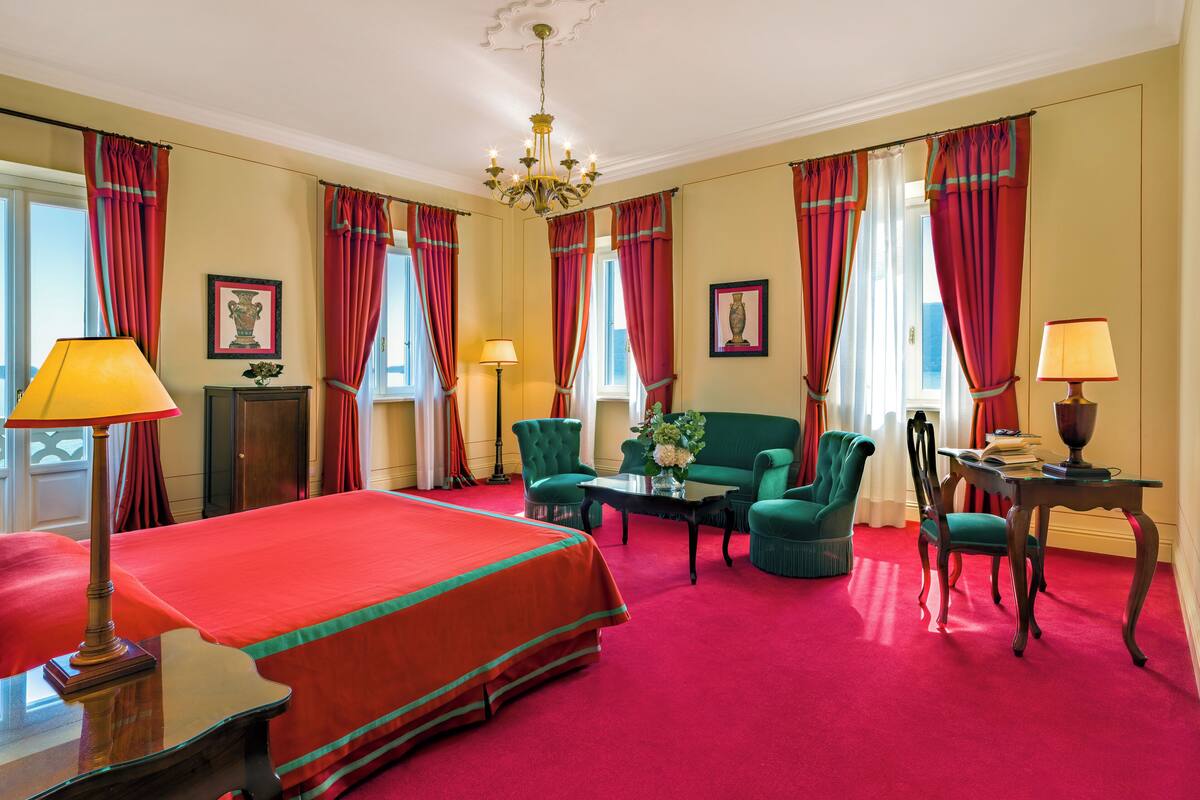 Een hotelkamer met een groot bed, twee fauteuils, een bank, een tafel met een lamp en uitzicht op zee.