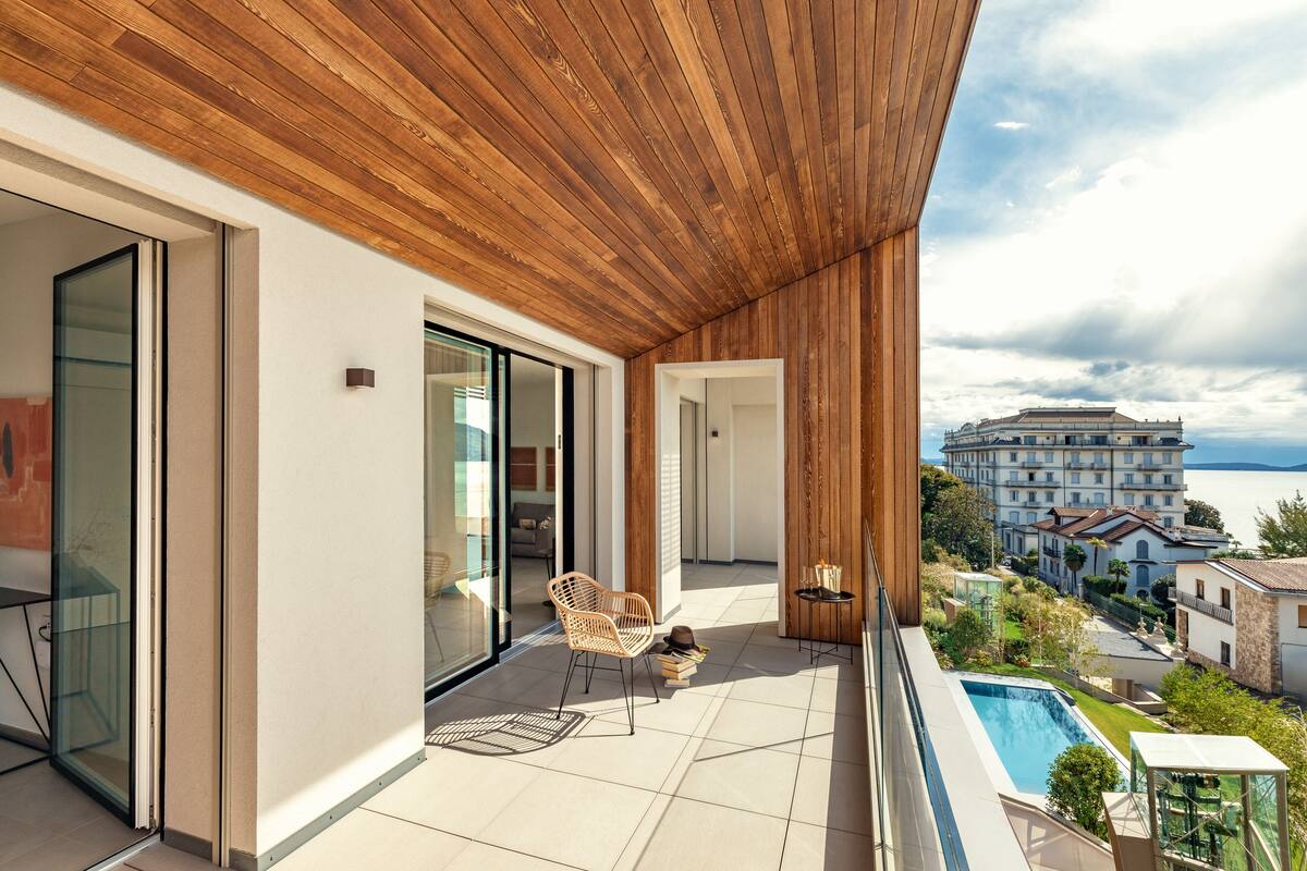 Een modern balkon met een houten plafond, een glazen deur die naar een kamer leidt, een rieten stoel, een klein tafeltje met boeken en een hoed, en een zwembadgedeelte met uitzicht op een groot gebouw en de zee.