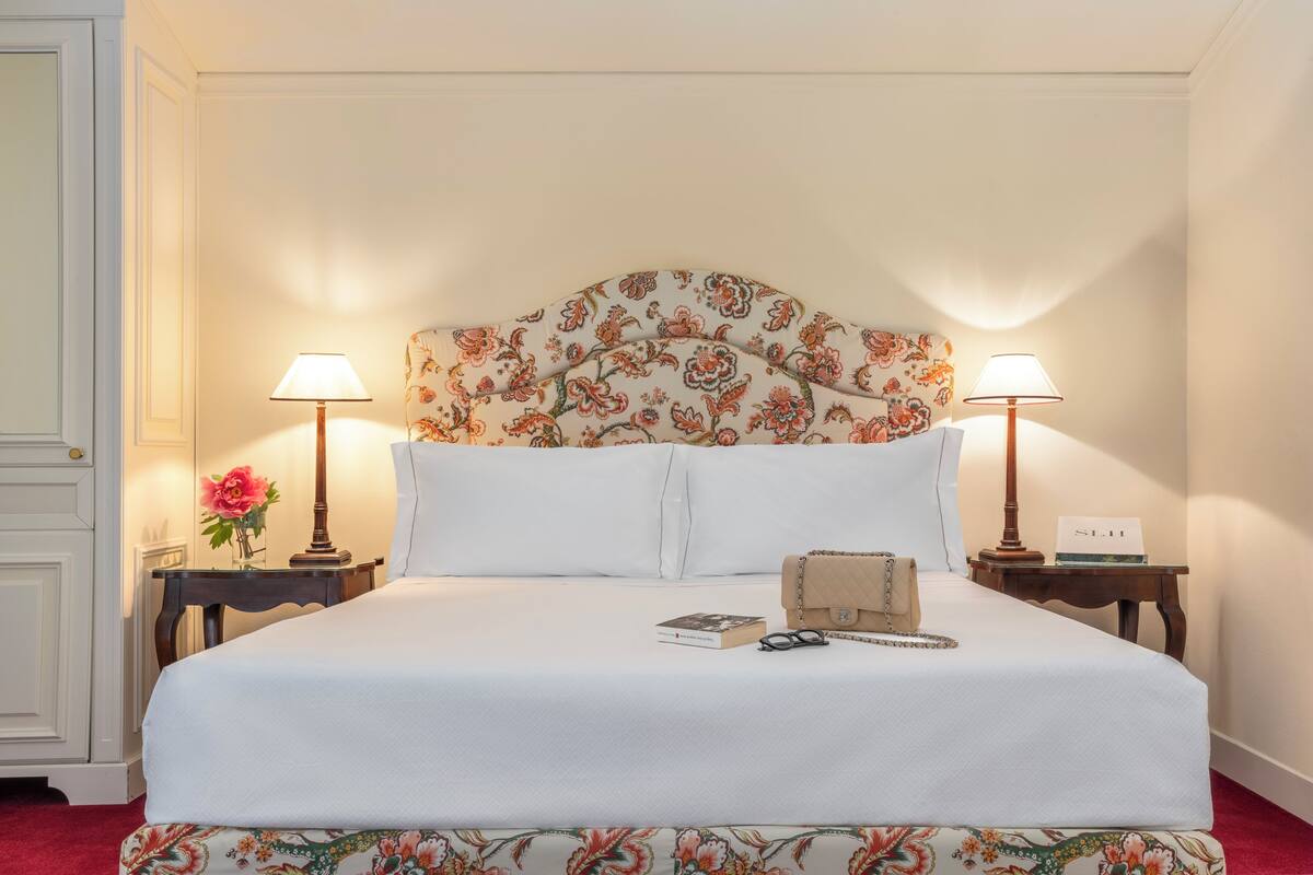 Een net opgemaakt bed met een bloemen hoofdbord, twee nachtkastjes met lampen, een klein nachtkastje met een boek, en een handtas op het bed.