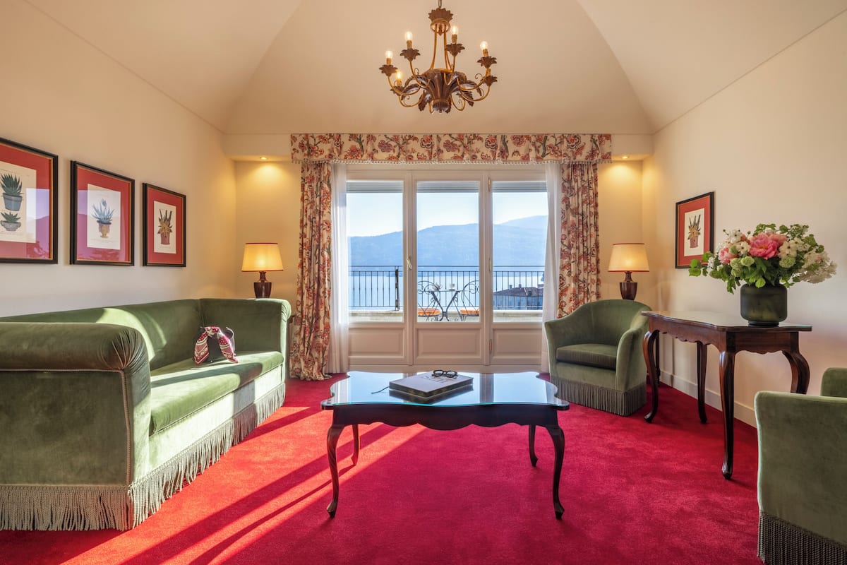 Een kamer met een groene bank, een bijpassende fauteuil, een glazen salontafel, een rood tapijt, een kroonluchter en uitzicht op zee.