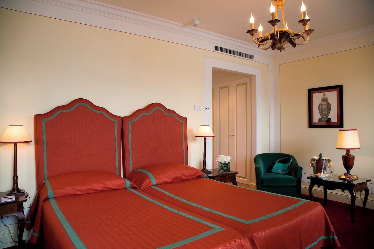 Een hotelkamer met een groot bed, twee nachtkastjes, een groene fauteuil, een kroonluchter en een ingelijst schilderij aan de muur.