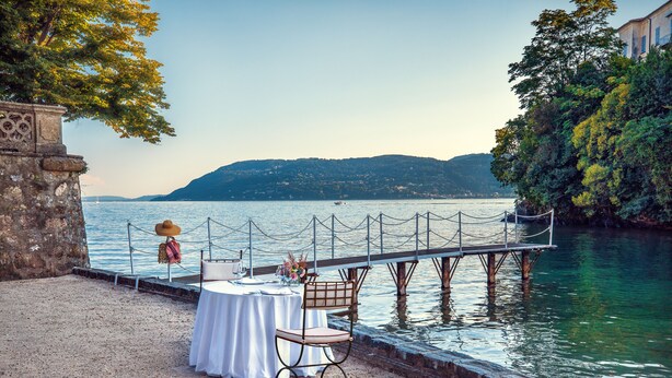 Une table dressée avec une nappe blanche, une chaise et un chapeau, le tout placé sur la rive d’un lac paisible, avec une jetée en bois qui s’avance dans l’eau.