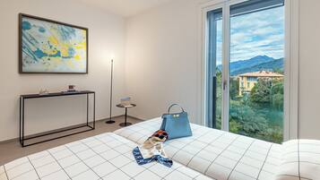 Une chambre d’hôtel moderne avec une grande fenêtre, un lit recouvert d’une courtepointe à motifs, un sac à main bleu posé sur le lit, un tableau abstrait encadré accroché au mur, et une petite table d’appoint avec une lampe et un livre.