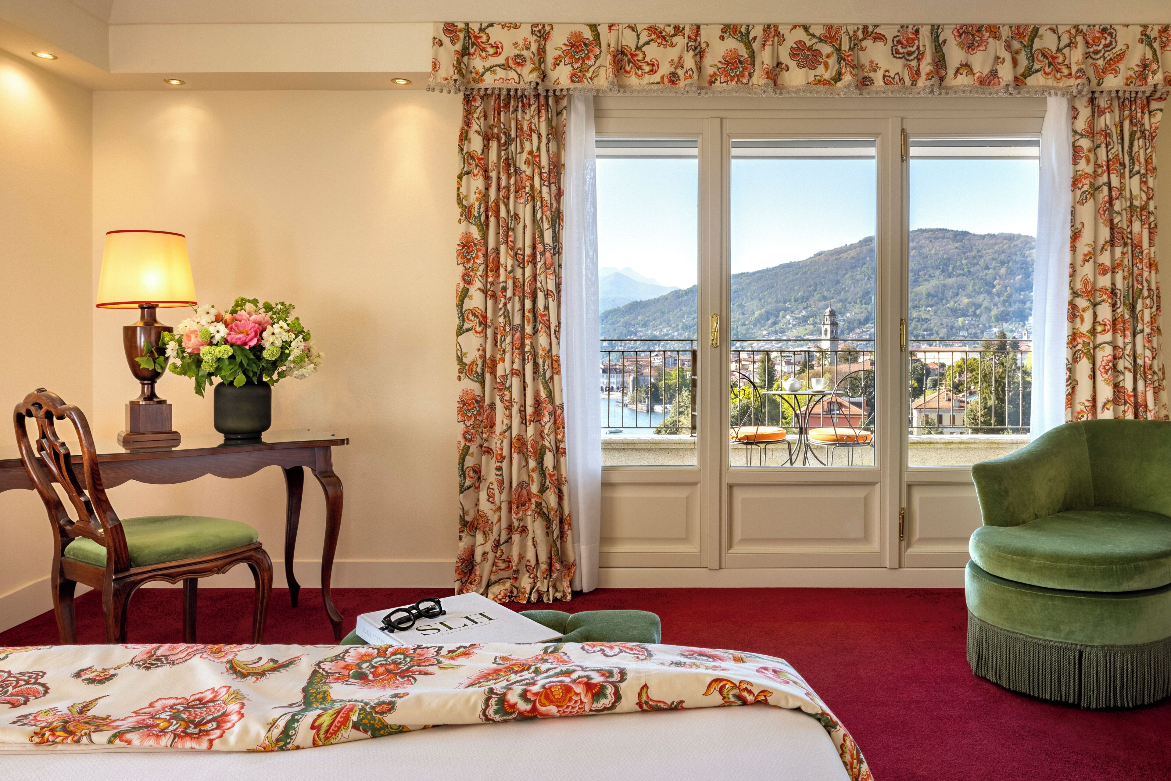 Une chambre d’hôtel avec un lit, un bureau avec une lampe, une chaise, un balcon avec vue sur les montagnes et un fauteuil vert.