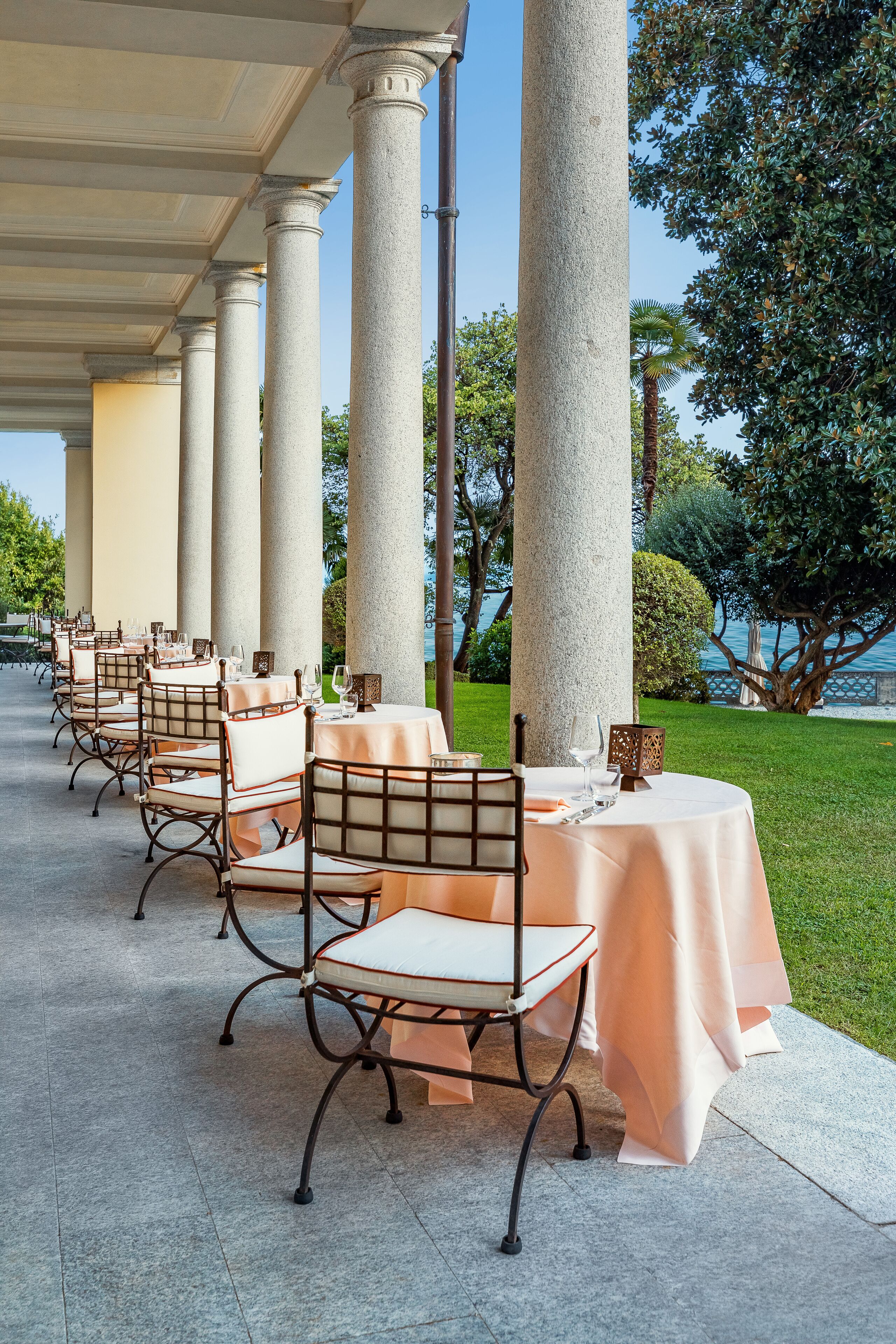 A covered outdoor dining area with tables and chairs, large columns, and a view of the sea.