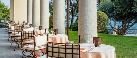 A covered outdoor dining area with tables and chairs, large columns, and a view of the sea.