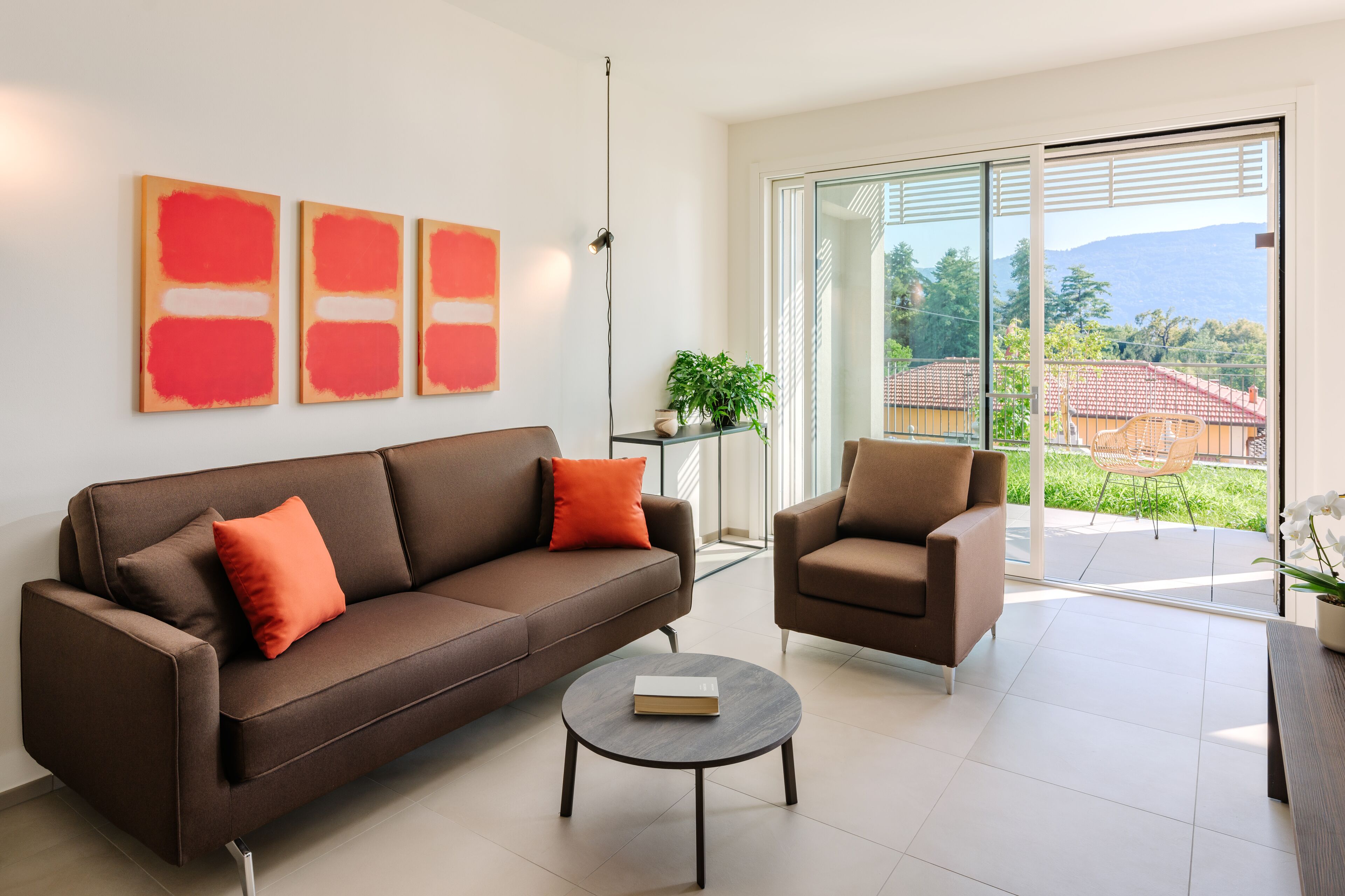 Ein modernes Wohnzimmer mit einem braunen Sofa, einem runden Couchtisch und einem braunen Sessel. An der Wand befinden sich drei abstrakte Gemälde und ein Blumentopf auf einem Tisch.